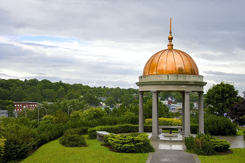 Photo of a structure with white columns and a copper bulb roof, surrounded by lush green hills.