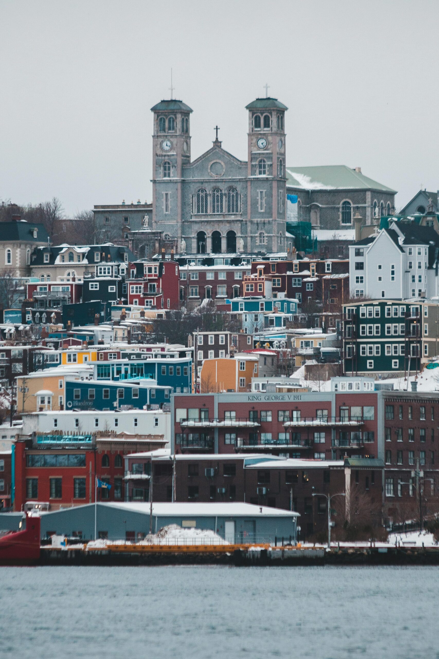 A photo of St. John's, Newfoundland and Labrador. There is a body of water at the bottom of the photo, then going upwards, there are rows of different coloured houses, and then at the top is a big church building.